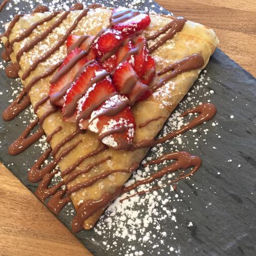 A delicious crepe topped with sliced strawberries, drizzled with chocolate, and sprinkled with powdered sugar on a slate plate.