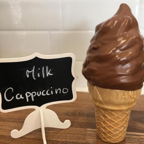 A chocolate soft-serve ice cream cone next to a small chalkboard sign that reads "Milk Cappuccino."