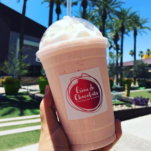 A hand holding a pink milkshake topped with whipped cream, set against a sunny outdoor backdrop with palm trees.