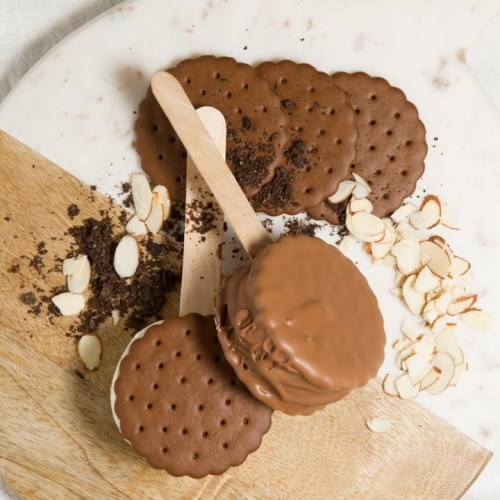 Chocolate ice cream sandwiches on a wooden board, with crushed cookies and sliced almonds scattered around.