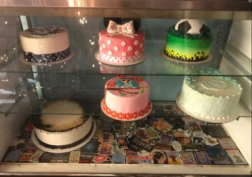 A display case with six colorful cakes, each decorated uniquely, on a background of various stickers and designs.