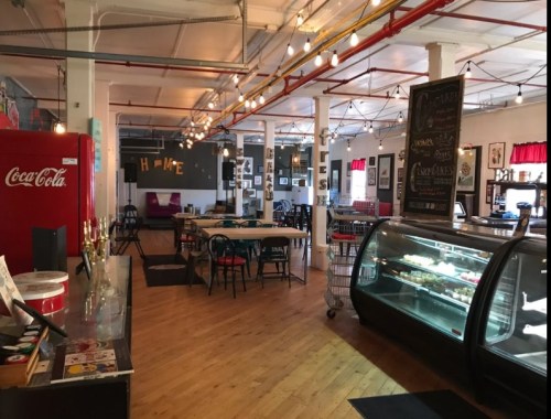 A cozy café interior with wooden floors, a Coca-Cola fridge, display case, and warm lighting. Art on the walls.