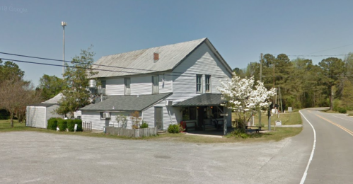 A weathered two-story building with a gray roof, surrounded by trees and a gravel road.