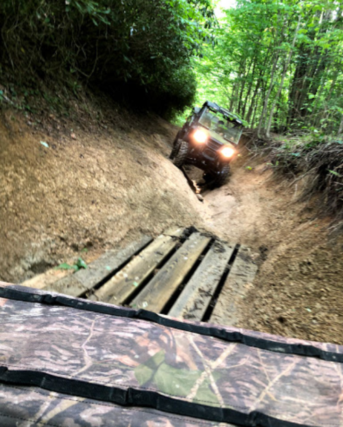 An off-road vehicle navigates a narrow, muddy trail with wooden planks, surrounded by dense greenery.