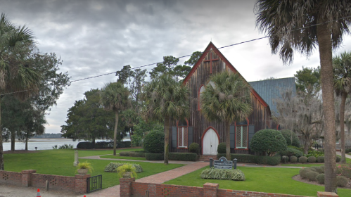 A rustic wooden church surrounded by lush greenery and palm trees, near a calm body of water under a cloudy sky.