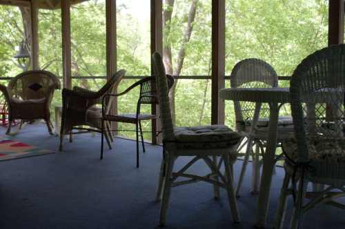 A screened porch with wicker chairs and a table, surrounded by greenery and natural light.