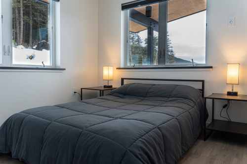 A cozy bedroom with a dark quilted bedspread, two lamps, and large windows overlooking a snowy landscape.