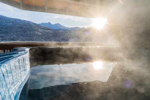 A serene hot tub with steam rising, surrounded by mountains and a bright sun shining in the background.