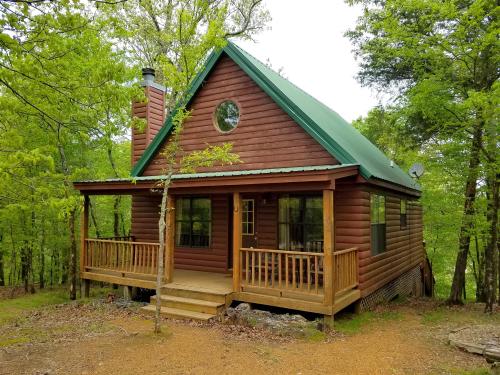 A rustic log cabin with a green metal roof, surrounded by trees and featuring a front porch and circular window.