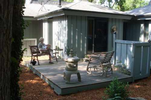 A serene outdoor deck with a woman reading, surrounded by greenery and a small stone table.