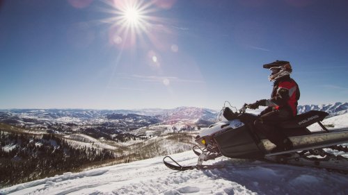 A snowmobiler sits on a snowy mountain, overlooking a vast landscape under a bright sun.
