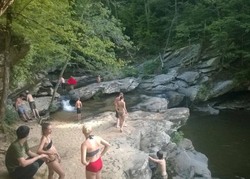 A group of people enjoying a natural swimming area with rocks and trees, some swimming and others relaxing by the water.