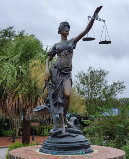 A bronze statue of Lady Justice holding scales and a sword, surrounded by greenery and palm trees.