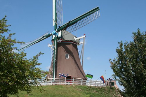 A large brown windmill with green blades, surrounded by trees and colorful flags, under a clear blue sky.