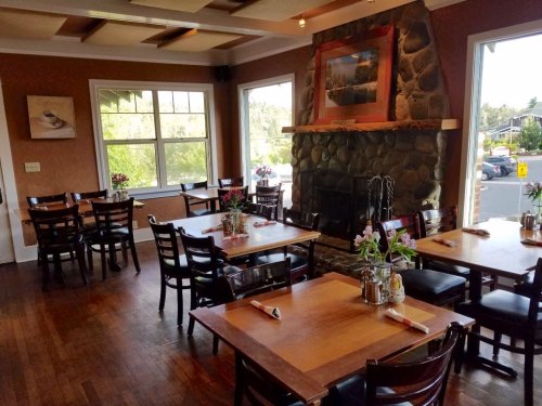 Cozy restaurant interior with wooden tables, chairs, and a stone fireplace, adorned with flowers and natural light.