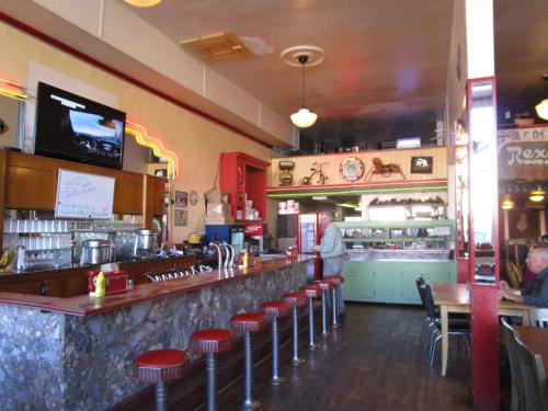 A retro diner interior with a long counter, red stools, and vintage decor, featuring a TV and a man at the counter.