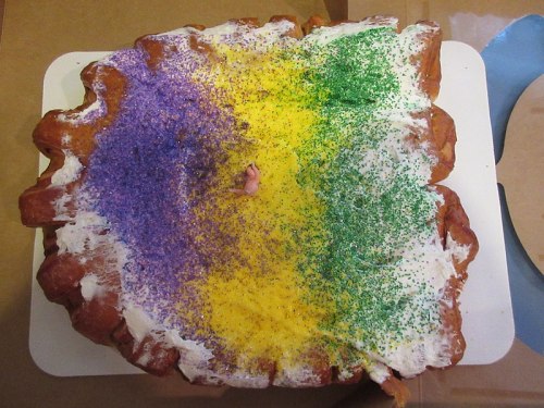 A colorful king cake topped with purple, yellow, and green sprinkles, featuring a small figurine in the center.