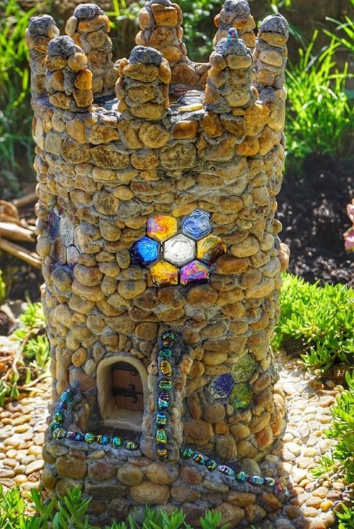 A whimsical stone tower with a colorful mosaic window, surrounded by greenery and pebbles.