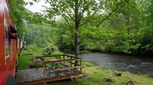A scenic view of a river beside a wooden deck, surrounded by lush green trees and a colorful train car.