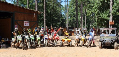 A group of motocross riders in gear poses with their bikes in a wooded area, celebrating with raised arms.
