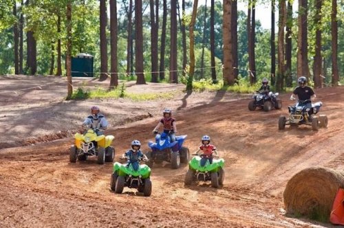 A group of children and adults riding ATVs on a dirt track surrounded by trees.