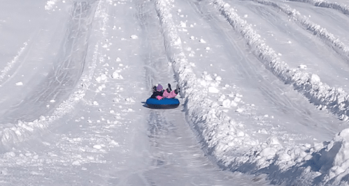 Two children in a blue snow tube slide down a snowy hill, surrounded by snowbanks and tracks in the snow.