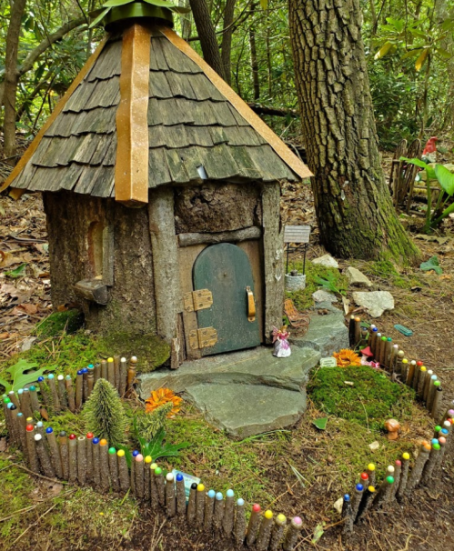 A whimsical fairy house made of wood and stones, surrounded by a colorful stick fence and lush greenery.