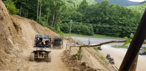 Two off-road vehicles navigate a dirt path on a hillside, surrounded by trees and mountains in the background.
