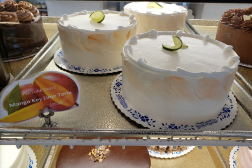 Two round cakes with white frosting, one labeled "Mango Key Lime Torte" priced at $39.00, displayed in a bakery case.