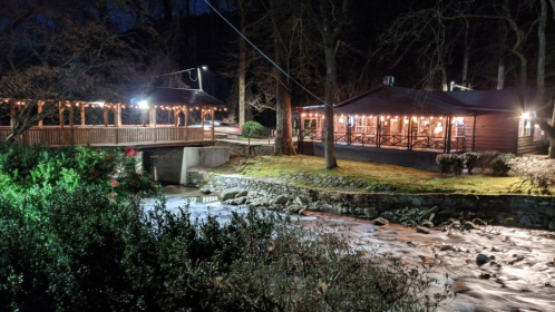 A cozy cabin by a stream at night, with warm lights and a bridge connecting to a nearby building.