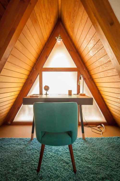 A cozy attic workspace featuring a wooden desk, a blue chair, and a triangular window with warm wooden walls.