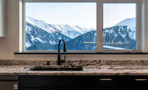 A modern kitchen sink with a black faucet, overlooking snow-capped mountains through a large window.