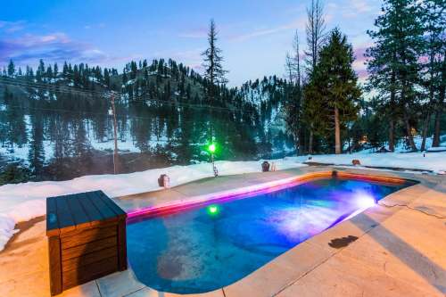 A scenic outdoor hot tub surrounded by snow-covered trees, with colorful lights and a mountain view at dusk.