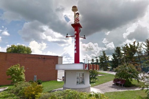 A tall, colorful sign for a museum, featuring a figure on top, surrounded by greenery and a cloudy sky.