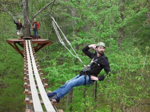 A person in a harness poses while ziplining through a lush green forest, with two others on a platform nearby.
