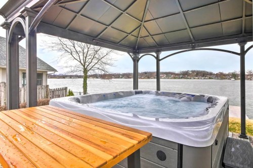A hot tub under a gazebo overlooking a calm lake, with a wooden table nearby and trees in the background.