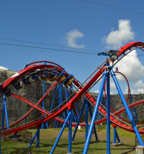 A colorful roller coaster with blue and red tracks against a clear blue sky, featuring a person sitting on top of a loop.