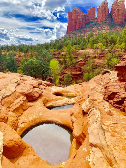 Rocky landscape with pools of water, surrounded by green trees and towering red rock formations under a cloudy sky.