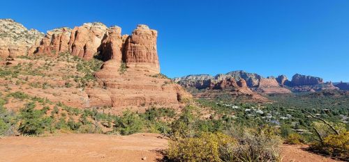 Red rock formations rise against a clear blue sky, with green vegetation and distant mountains in the background.