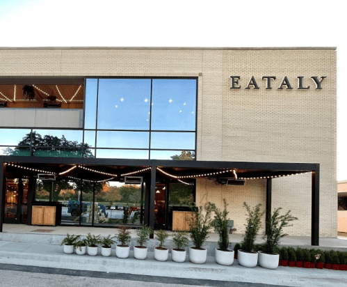 Exterior of Eataly building with large windows, string lights, and potted plants in front.