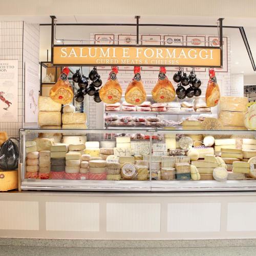 A display of cured meats and cheeses, featuring hanging hams and various cheese wheels in a market setting.