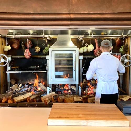 A chef in a white coat cooks over an open flame in a rustic kitchen with hanging meats and vegetables.