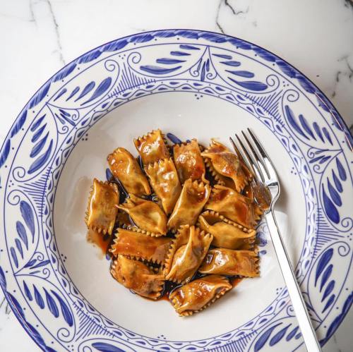 A decorative plate filled with ravioli drizzled in sauce, accompanied by a silver fork on a marble surface.