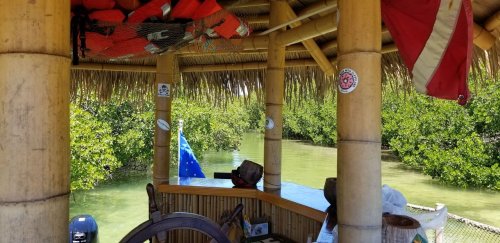 A bamboo hut overlooking a calm waterway, surrounded by lush greenery and decorated with flags and stickers.
