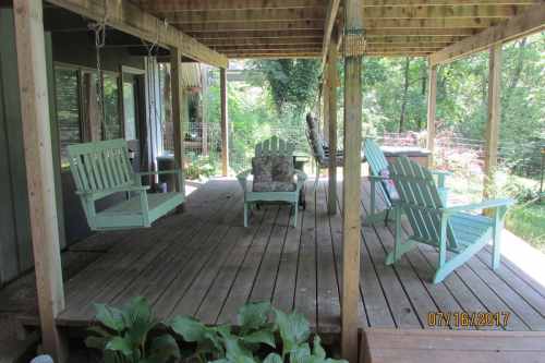 A wooden porch with green adirondack chairs and swings, surrounded by greenery and trees.