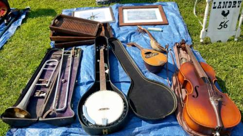 A collection of musical instruments and framed art displayed on a blue tarp, including a banjo, violin, and trumpet.