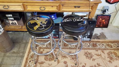 Two bar stools with black seats featuring Elvis Presley signatures, placed in a room with wooden furniture and decor.