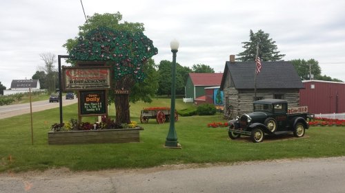 A rustic restaurant sign with a tree design, an old car, and a charming building surrounded by flowers and greenery.