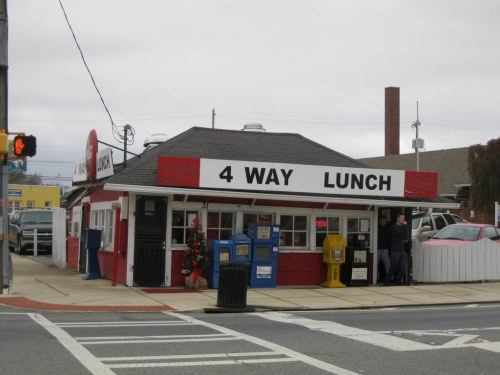 A small diner named "4 Way Lunch" with a red exterior, featuring a sign and nearby newspaper boxes.