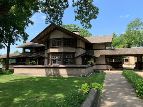 A large, two-story house with a unique architectural design, surrounded by green grass and trees under a clear blue sky.